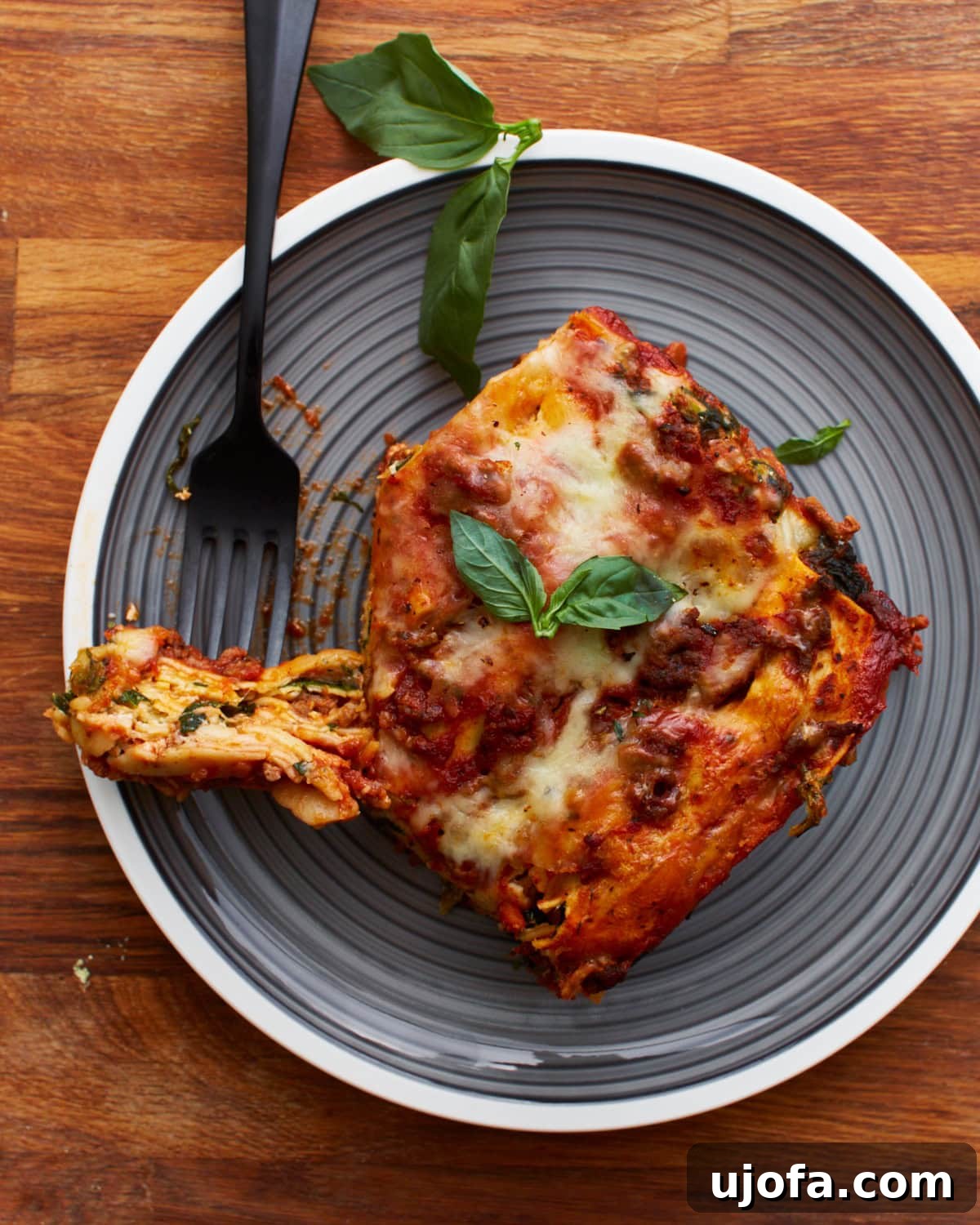 Irresistible Gluten-Free Lasagna 16 A black fork holding a piece of gluten-free lasagna on a grey plate, garnished with fresh basil leaves, highlighting its appetizing layers.