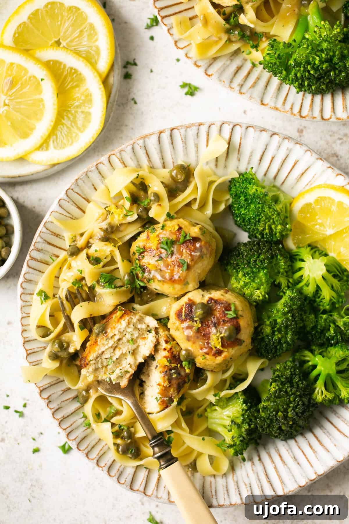 Zesty Gluten-Free Chicken Piccata Meatballs 2 chicken piccata meatballs with gluten free pasta and broccoli on a white scalloped plate with parsley and lemon