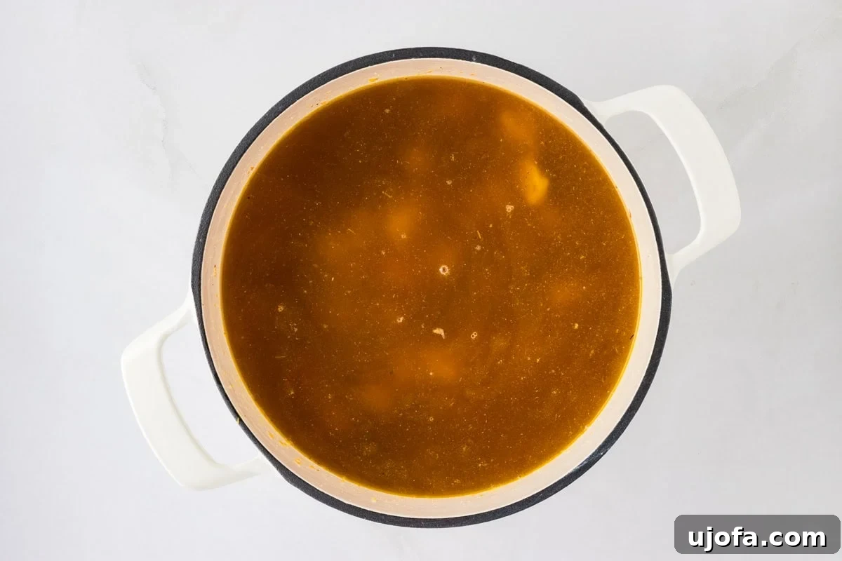 A large white pot filled with golden chicken broth, simmering gently on a stovetop. The broth is clear, with flecks of seasoning visible, signifying it's ready for the next ingredient.