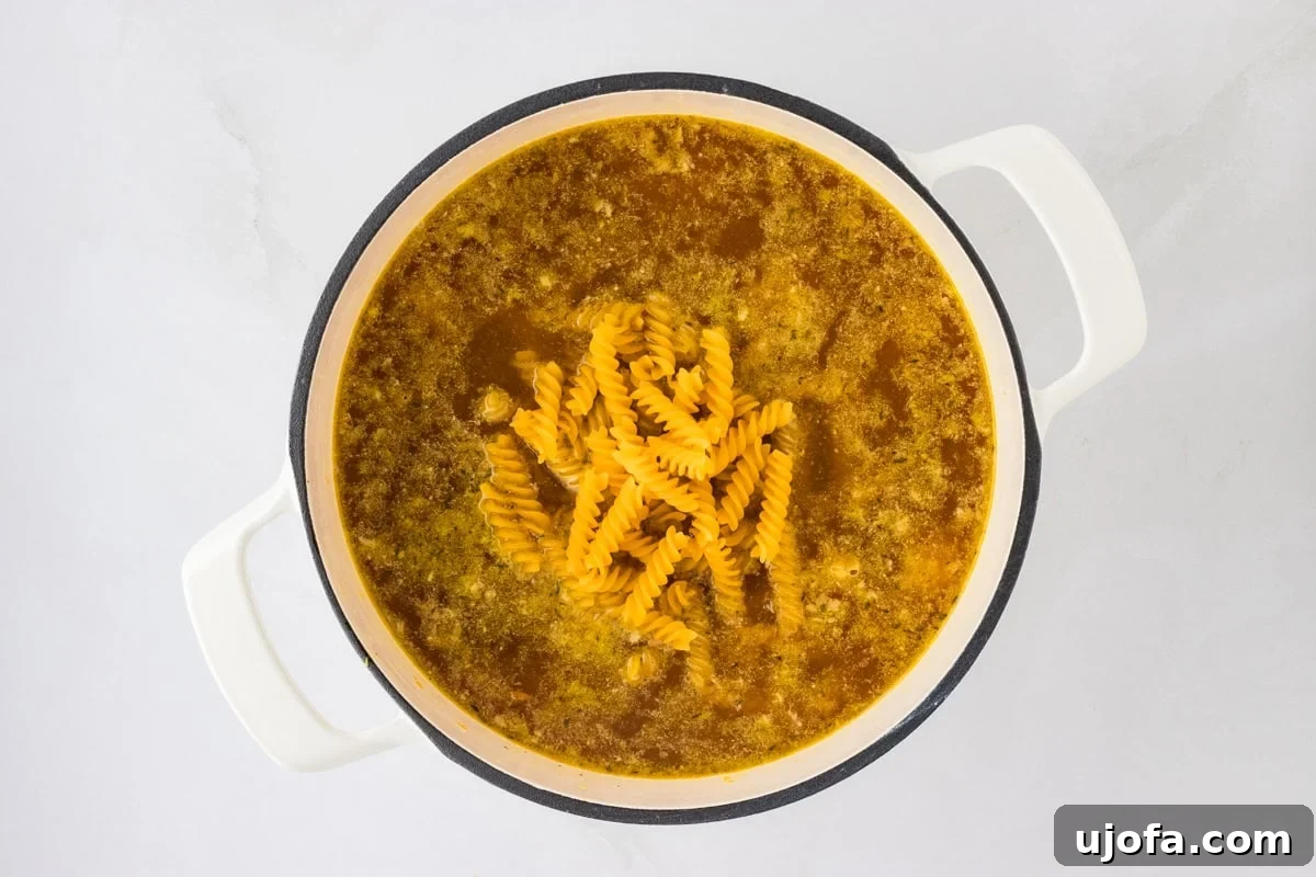 Spiral-shaped gluten-free noodles are being added into a pot of simmering chicken broth and vegetables, illustrating the stage where the pasta is introduced to the soup.