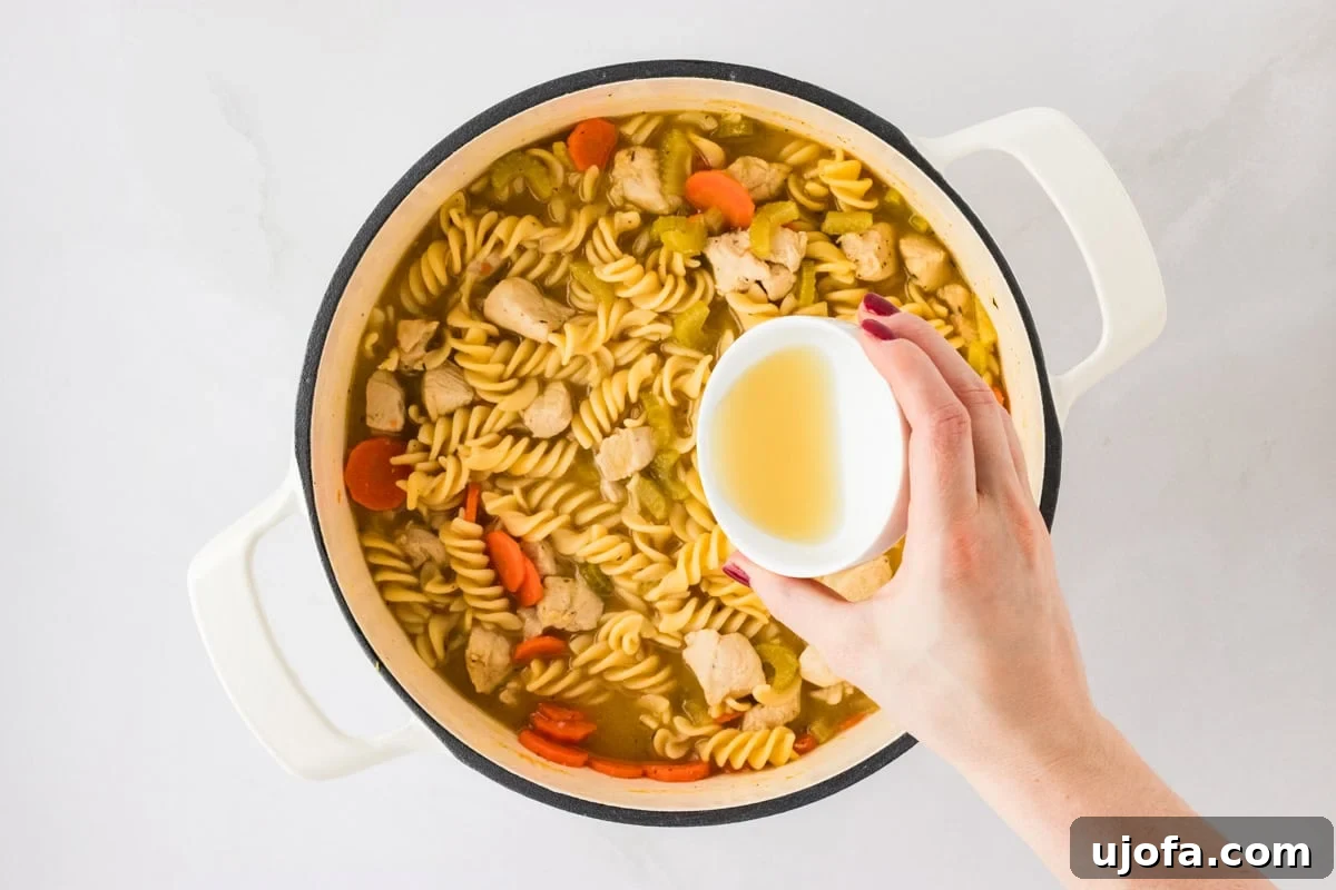 A pot of vibrant, fully cooked gluten-free chicken noodle soup, with spiral noodles, tender chicken chunks, carrots, and celery visible. A hand is shown adding a splash of liquid, likely lemon juice or apple cider vinegar, to enhance the flavor.