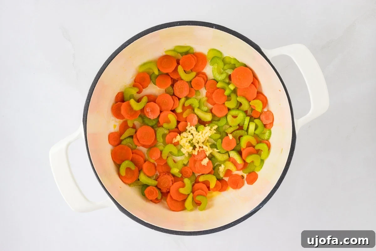 A close-up inside a white pot, showing sautéed carrots and celery blended with minced garlic. The vegetables are lightly softened, and the garlic is fragrant, indicating the next stage of soup preparation.