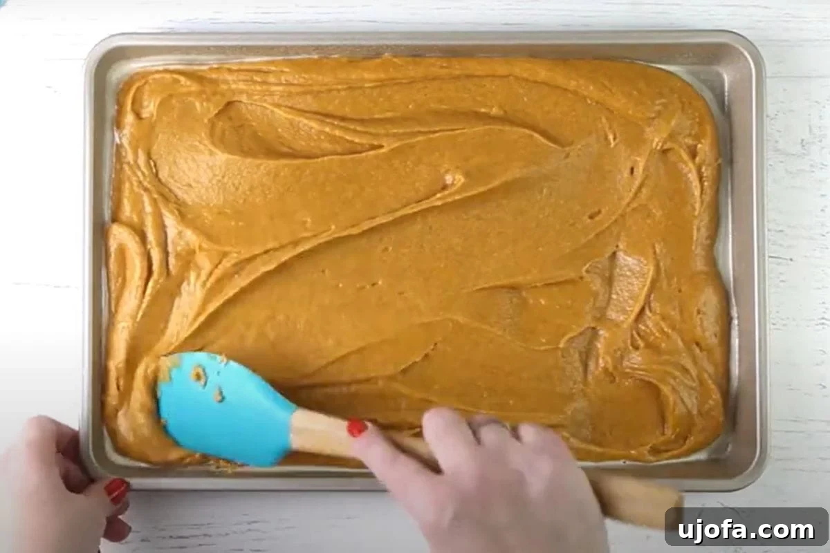 Decadent Gluten-Free Pumpkin Spice Squares 11 A hand gently uses a spatula to smooth out the rich, orange gluten-free pumpkin bar batter evenly across a parchment-lined baking tray, ensuring a uniform layer before it goes into the oven.