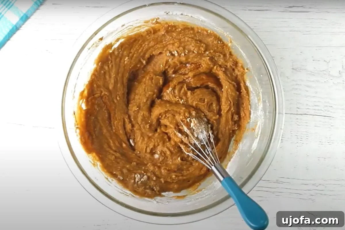 Decadent Gluten-Free Pumpkin Spice Squares 9 The fluffy, light brown gluten-free flour has been carefully added to the pumpkin, sugar, and egg batter in a large glass mixing bowl. The mixture is now thicker, transitioning from a liquid to a more substantial dough, ready for its final ingredients.