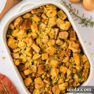 Savory Gluten-Free Cornbread Dressing 3 A baked dish of golden-brown gluten-free cornbread stuffing with herbs, served in a white casserole dish with a wooden spoon and fresh rosemary nearby.