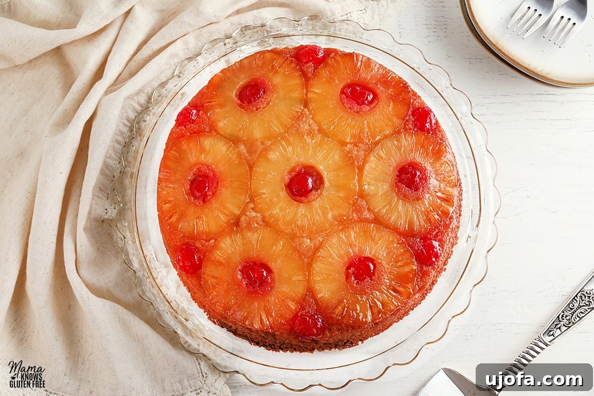 A vibrant gluten-free pineapple upside-down cake, beautifully displayed on a glass cake plate, with a cake cutter and plates ready in the background.