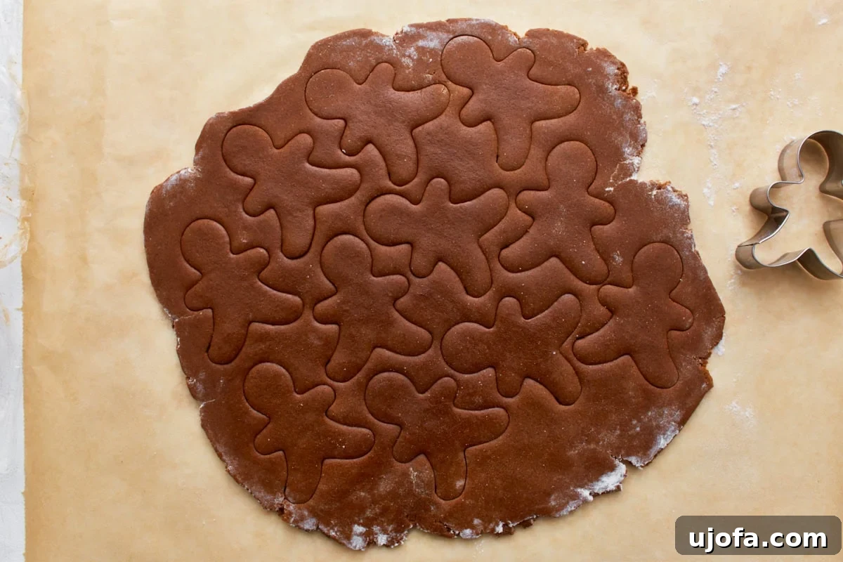 Guilt-Free Gingerbread Bliss 12 Gluten-free gingerbread dough, rolled out evenly on parchment paper, with several gingerbread man shapes already cut out and resting, alongside a cookie cutter.