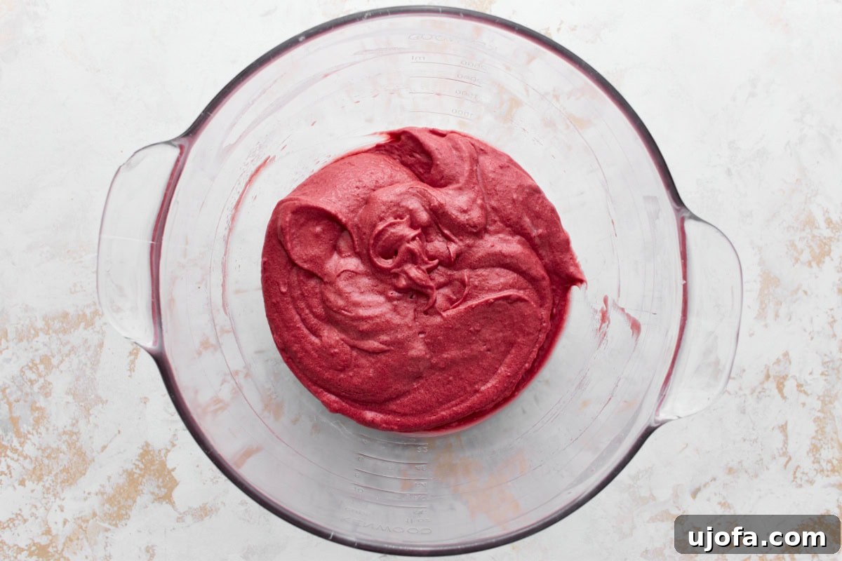 A glass mixing bowl containing smooth gluten-free red velvet cake batter, sitting on a textured white and beige surface.