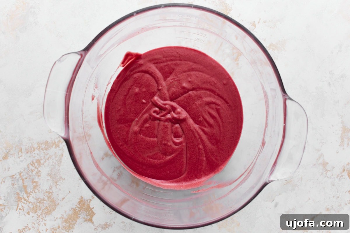 A glass mixing bowl containing gluten free red velvet cake batter sitting on a textured white and beige surface.