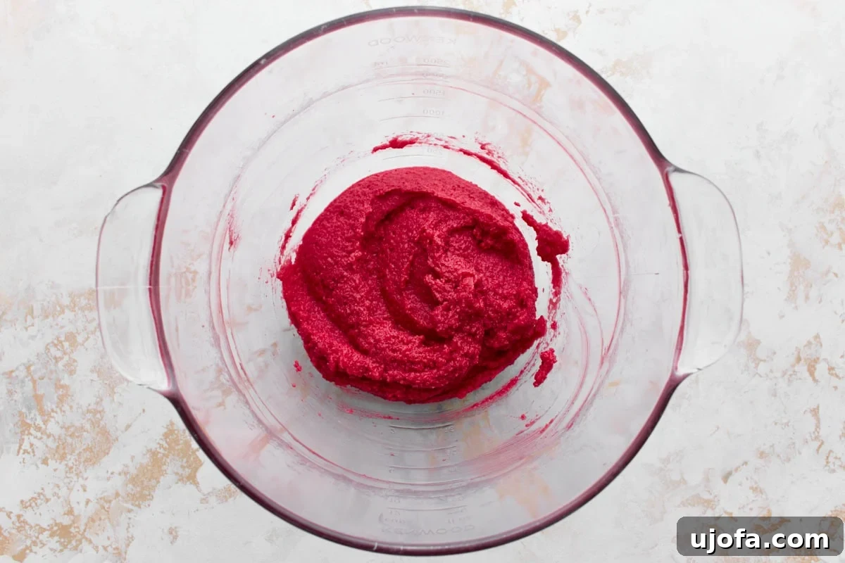 A glass mixing bowl containing vibrant red creamed butter, thick and smooth in texture, sitting on a textured white and beige surface.