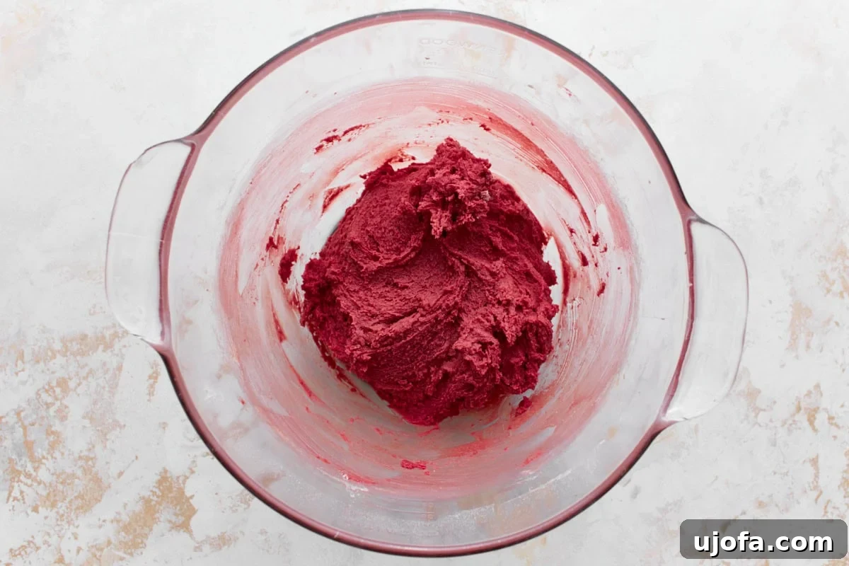 A glass mixing bowl containing vibrant red cake batter, thick and smooth in texture, sitting on a textured white and beige surface.