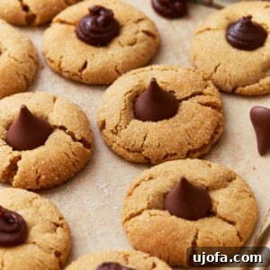 A festive tray of gluten-free peanut butter blossom cookies, featuring golden, crackled edges and classic Hershey's Kisses pressed into their centers, all resting on parchment paper.