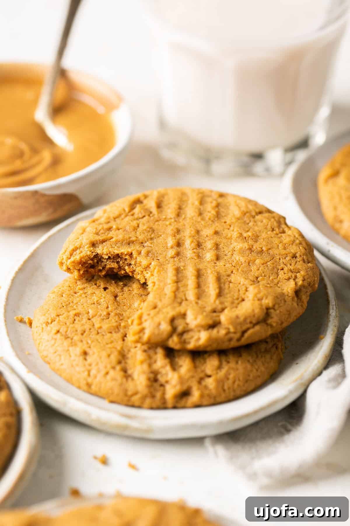 Two soft gluten-free peanut butter cookies stacked on a small white plate, with a background blur of a bowl of creamy peanut butter.