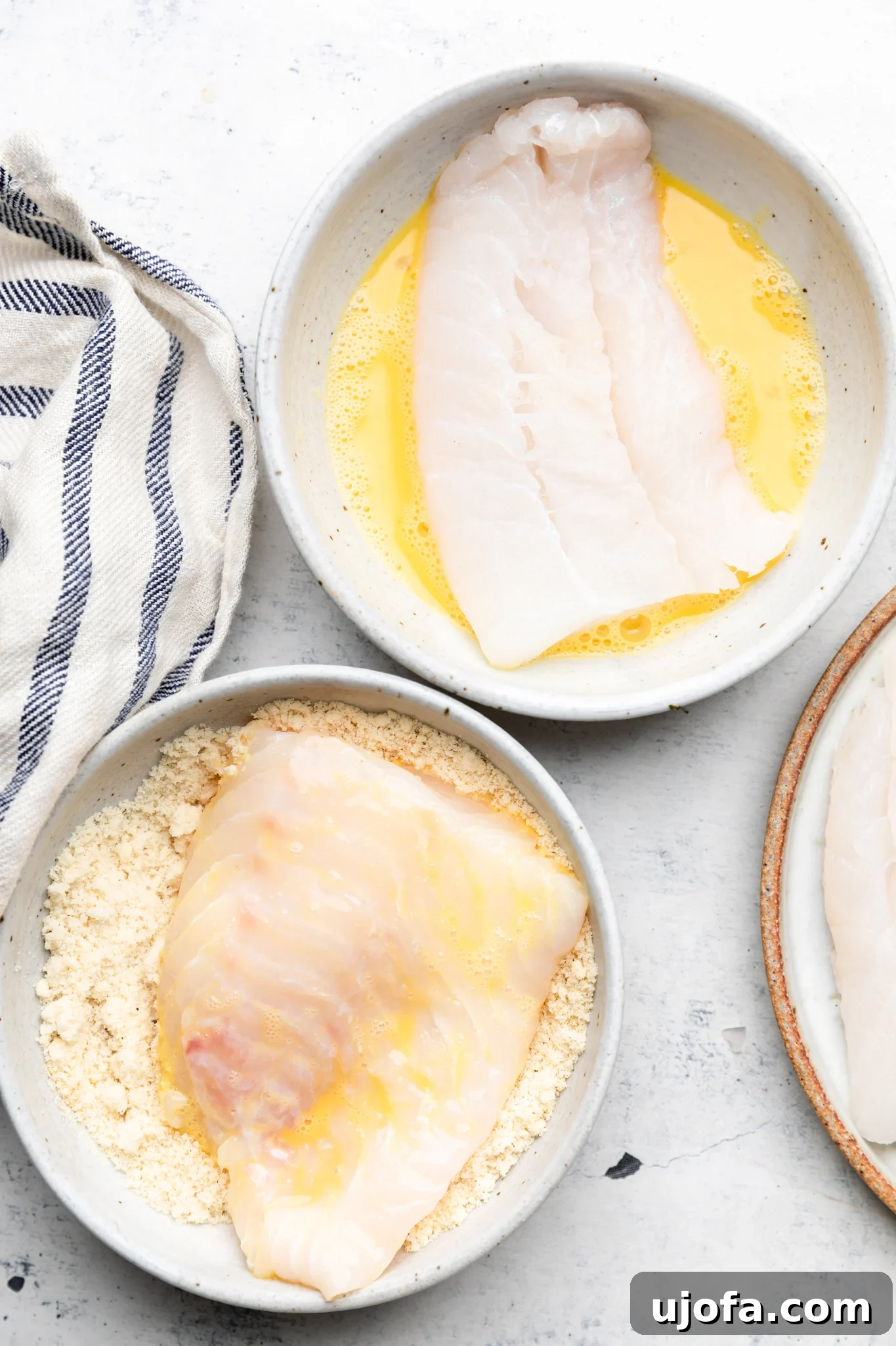 Close-up of cod fillets being dipped first in an egg wash and then into almond flour, preparing them for cooking.