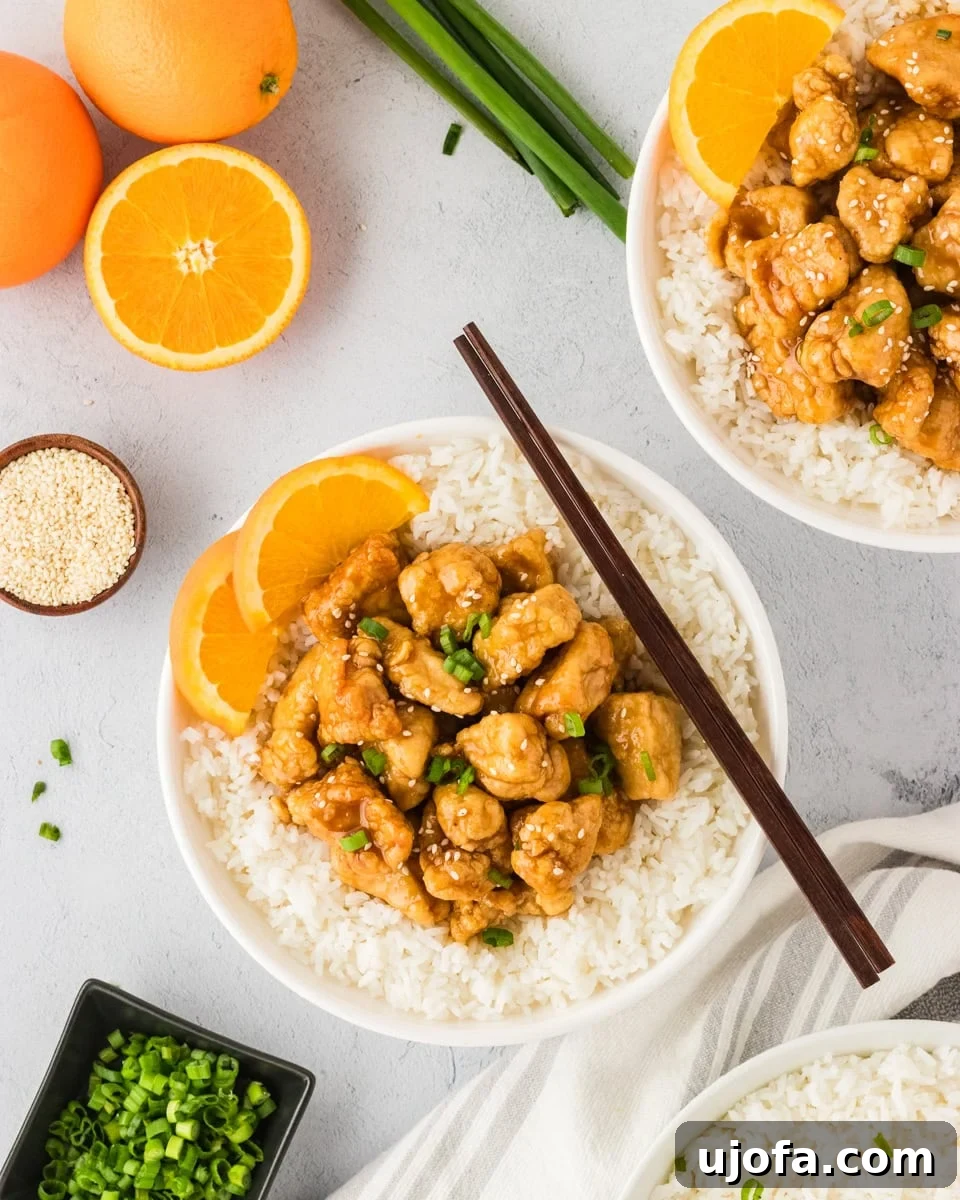 Tangy Gluten-Free Orange Chicken 2 A white bowl filled with gluten-free orange chicken served over white rice, garnished with fresh orange slices, green onions, and sesame seeds. Whole oranges and scallions are displayed in the background for a fresh presentation.