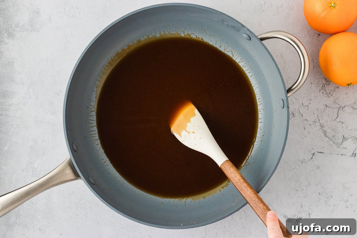 Tangy Gluten-Free Orange Chicken 16 A skillet of glossy, thickened orange sauce being stirred with a white spatula, surrounded by fresh oranges.