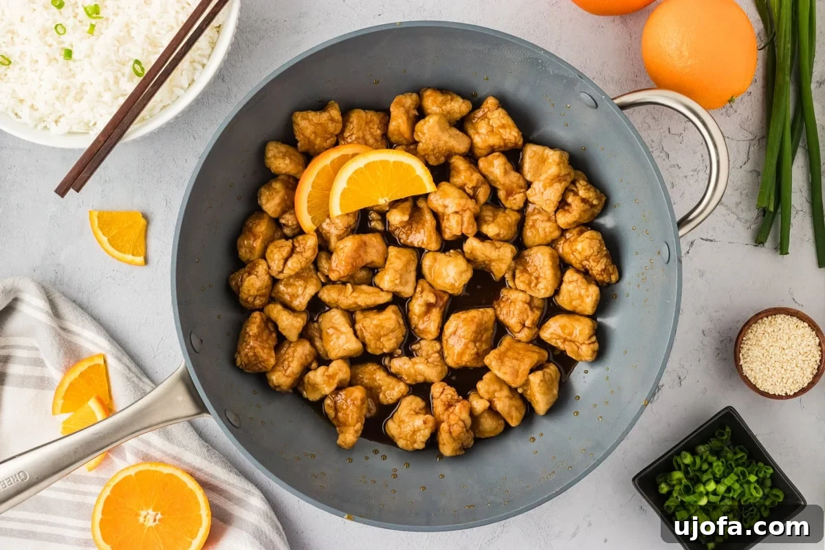 Tangy Gluten-Free Orange Chicken 18 A skillet of gluten-free orange chicken coated in glossy sauce, garnished with orange slices, surrounded by bowls of rice, sesame seeds, and scallions for serving.