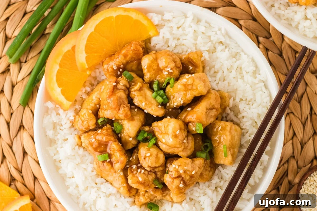 Tangy Gluten-Free Orange Chicken 19 A close-up of a bowl of gluten-free orange chicken served over fluffy white rice, garnished with sesame seeds, green onions, and fresh orange slices, with chopsticks resting on the side.