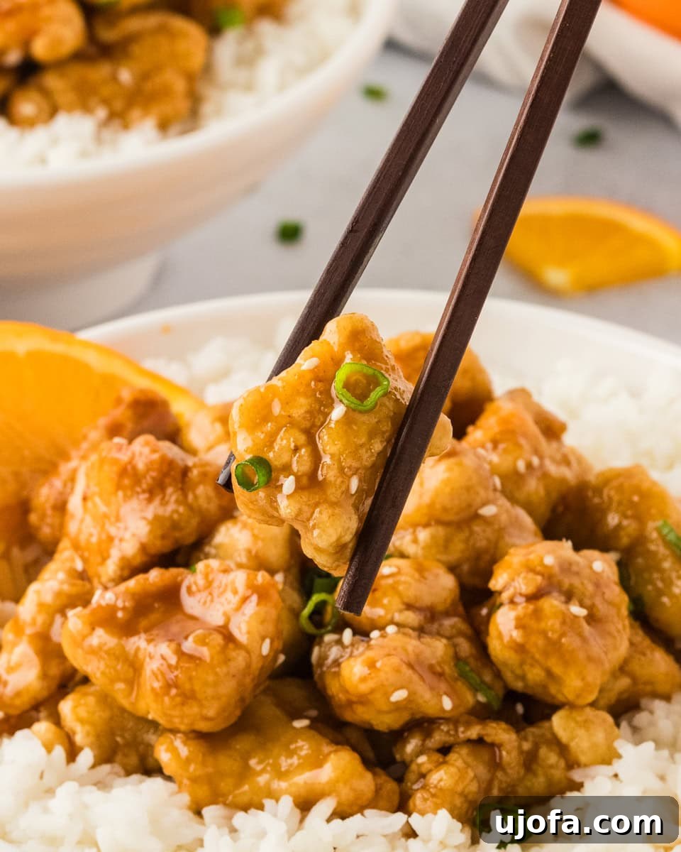 Tangy Gluten-Free Orange Chicken 20 A close-up of chopsticks holding a piece of gluten-free orange chicken with glossy sauce, garnished with green onions and sesame seeds, with more chicken and rice visible in the background.