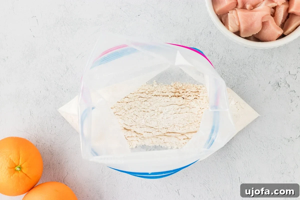 Tangy Gluten-Free Orange Chicken 5 A Ziploc bag containing gluten-free flour mixture for breading chicken, with fresh oranges and diced raw chicken visible on a white countertop.