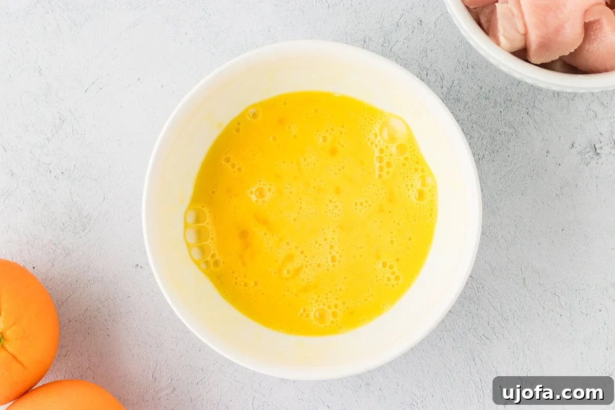 Tangy Gluten-Free Orange Chicken 6 A bowl of whisked eggs ready for dipping chicken pieces, surrounded by fresh oranges and diced raw chicken on a textured countertop.
