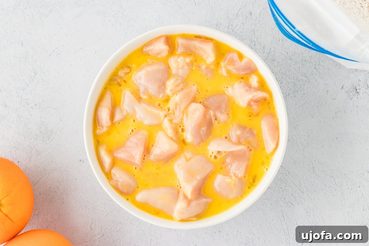 Tangy Gluten-Free Orange Chicken 7 Raw chicken pieces coated in egg in a white bowl, with oranges and flour mixture visible nearby, ready for breading.