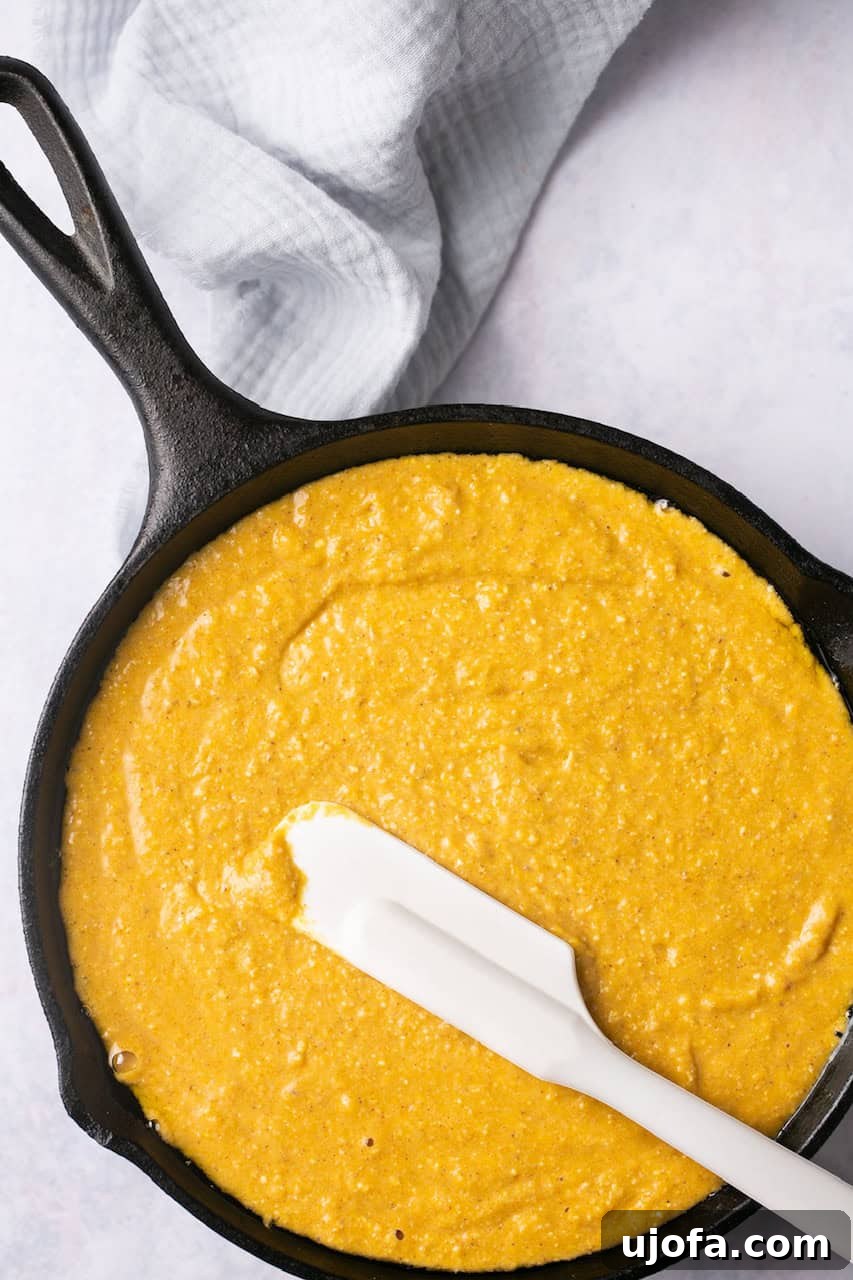 Unbaked sweet potato cornbread batter resting in a cast iron pan with a white spatula placed on top, ready for the oven.