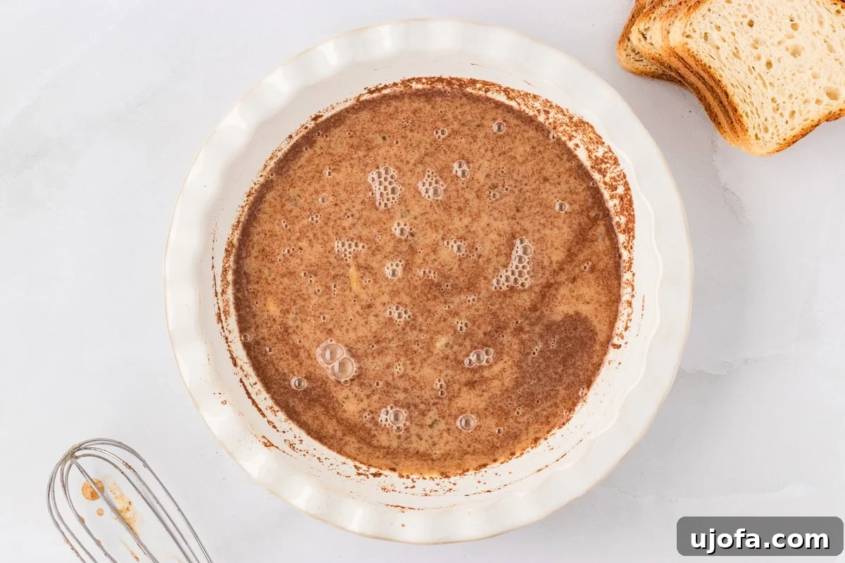 Overhead view of a shallow dish filled with whisked egg mixture spiced with cinnamon and nutmeg, with slices of gluten-free bread beside it.