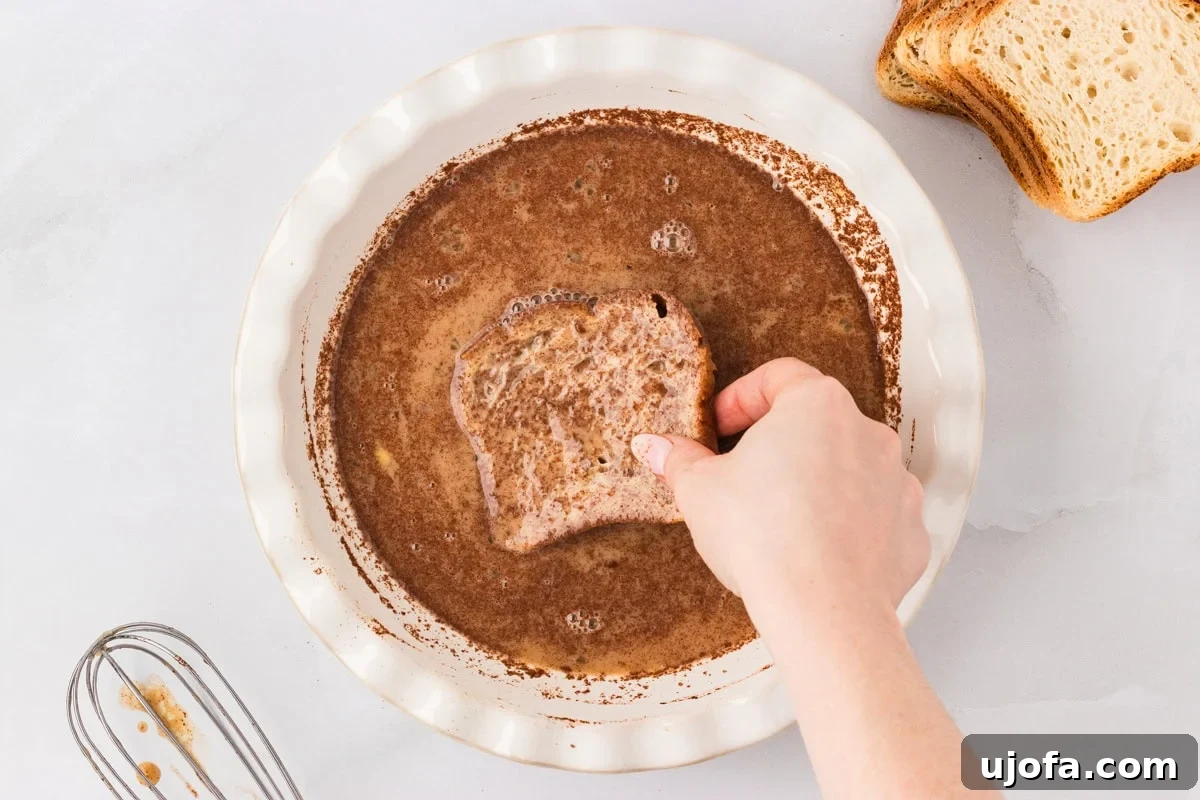 A hand dipping a slice of gluten-free bread into the spiced egg mixture in a pie dish, with other slices of bread on the side.
