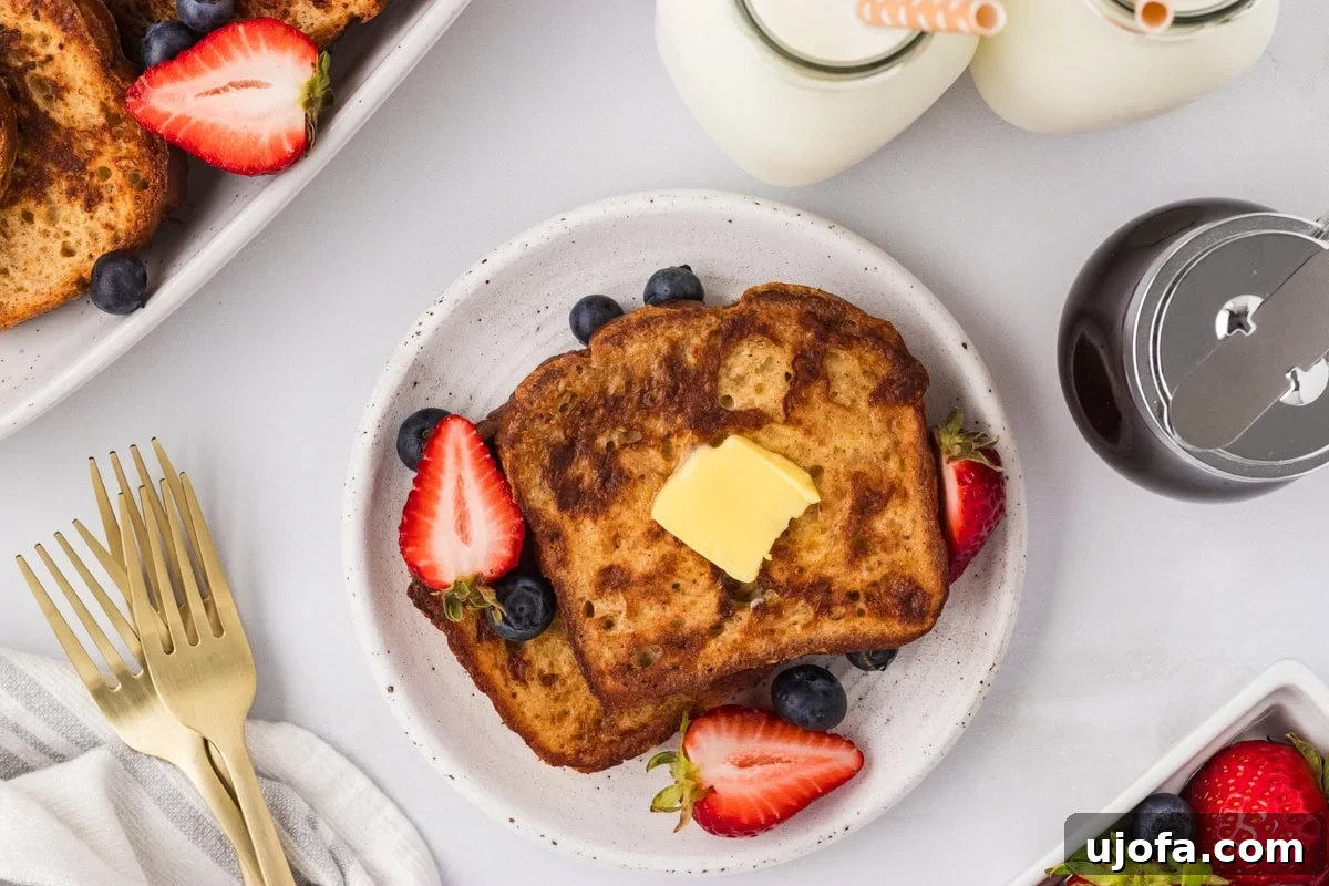 Plate of cooked gluten-free French toast topped with butter and surrounded by fresh strawberries and blueberries, with milk and syrup in the background.