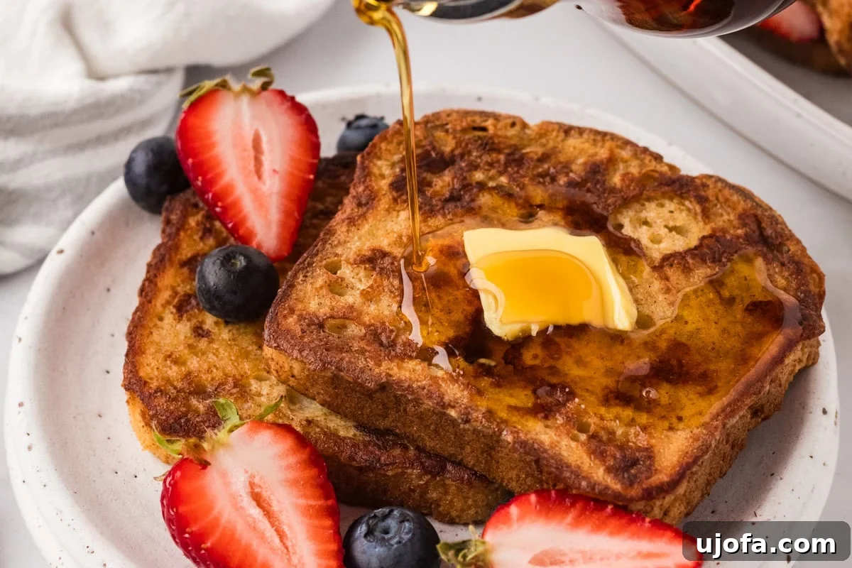 Close-up of French toast on a plate with a pat of melting butter and syrup being poured over the top, served with fresh berries.