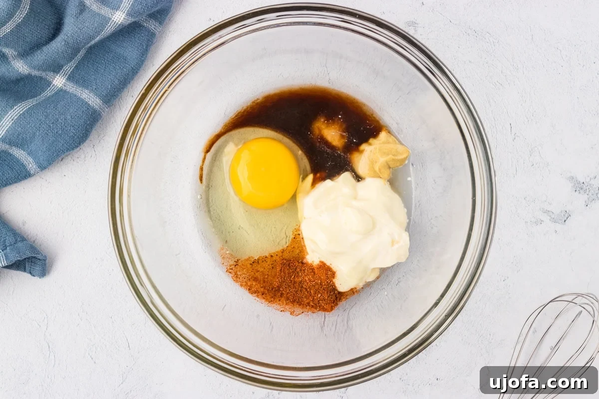 A glass mixing bowl with egg, mayonnaise, Dijon mustard, Worcestershire sauce, and seasonings ready to be whisked together into a smooth wet mixture.