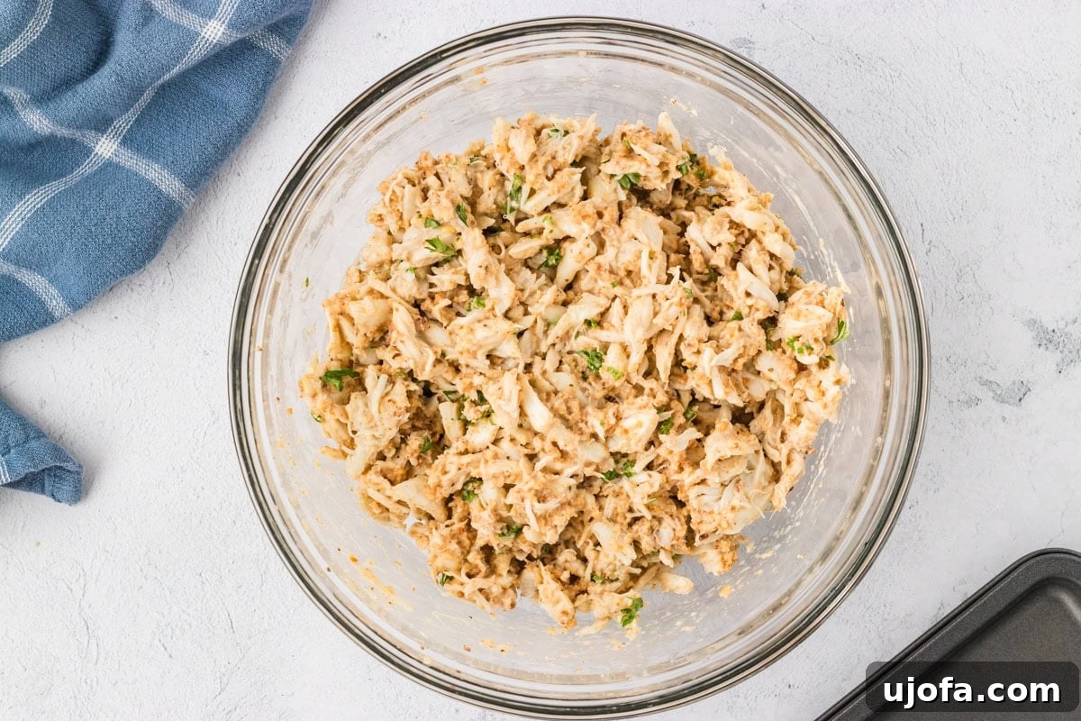 Crab cake mixture fully combined in a glass bowl, showing large chunks of crab and flecks of parsley, covered with plastic wrap, ready for chilling.