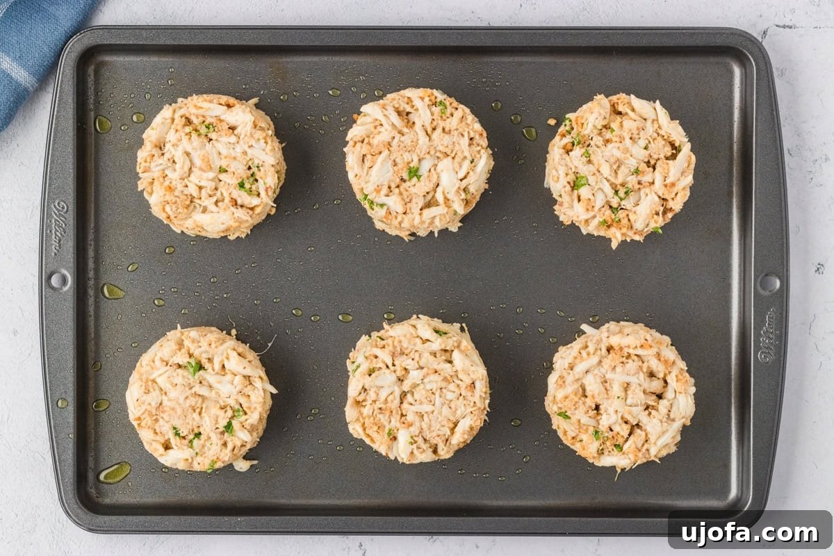 Six unbaked crab cakes formed into perfect mounds on an oiled baking sheet, evenly spaced and ready to cook.