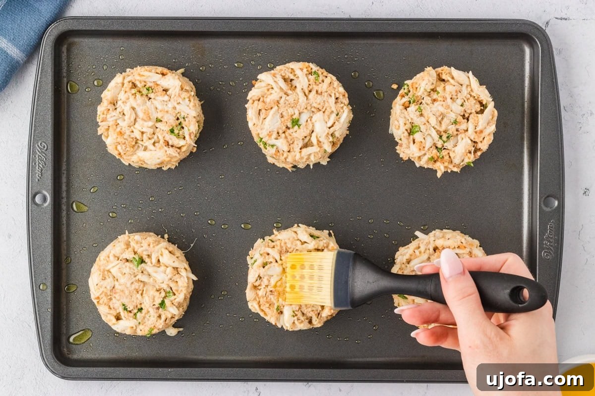 A hand brushing the tops of unbaked crab cakes with melted butter on a baking sheet, preparing them for baking.