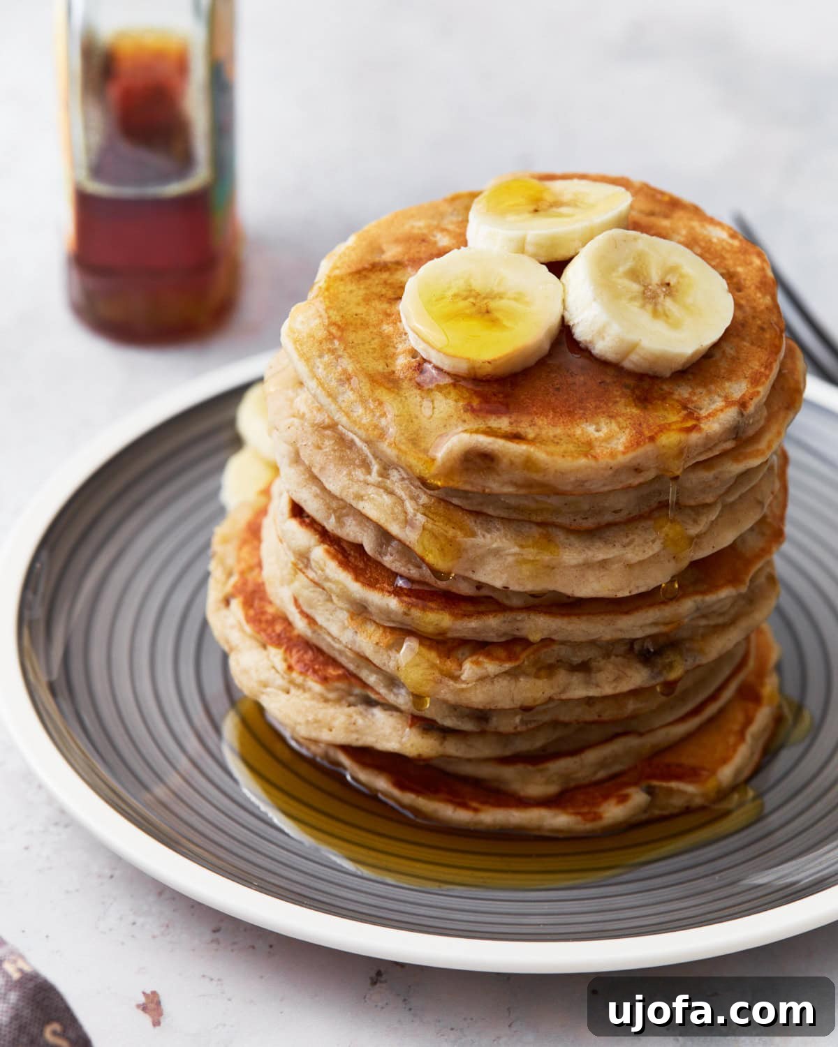 A stack of golden-brown gluten-free banana pancakes on a gray plate, artfully topped with fresh banana slices and drizzled with glistening maple syrup. A bottle of syrup stands blurred in the warm background.