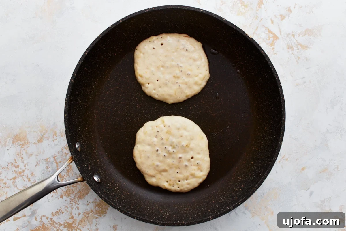 Two golden-brown gluten-free banana pancakes cooking on a non-stick skillet, with distinct bubbles forming on the surface, a clear sign they are ready to be flipped.