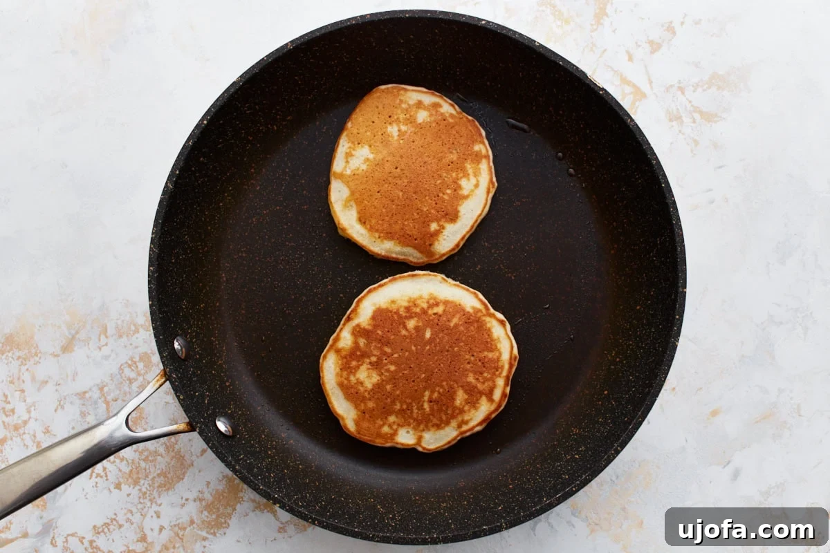 Two perfectly cooked, golden-brown gluten-free banana pancakes in a non-stick skillet, having been freshly flipped and ready to be served, highlighting their inviting texture.