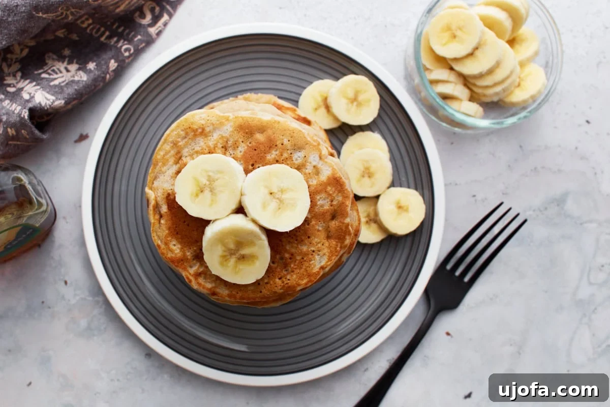 A delightful top-down view of a generous stack of gluten-free banana pancakes nestled on a gray plate, elegantly garnished with fresh banana slices. Adjacent to the stack, a small glass bowl overflows with more banana slices, and a bottle of maple syrup stands poised for drizzling.