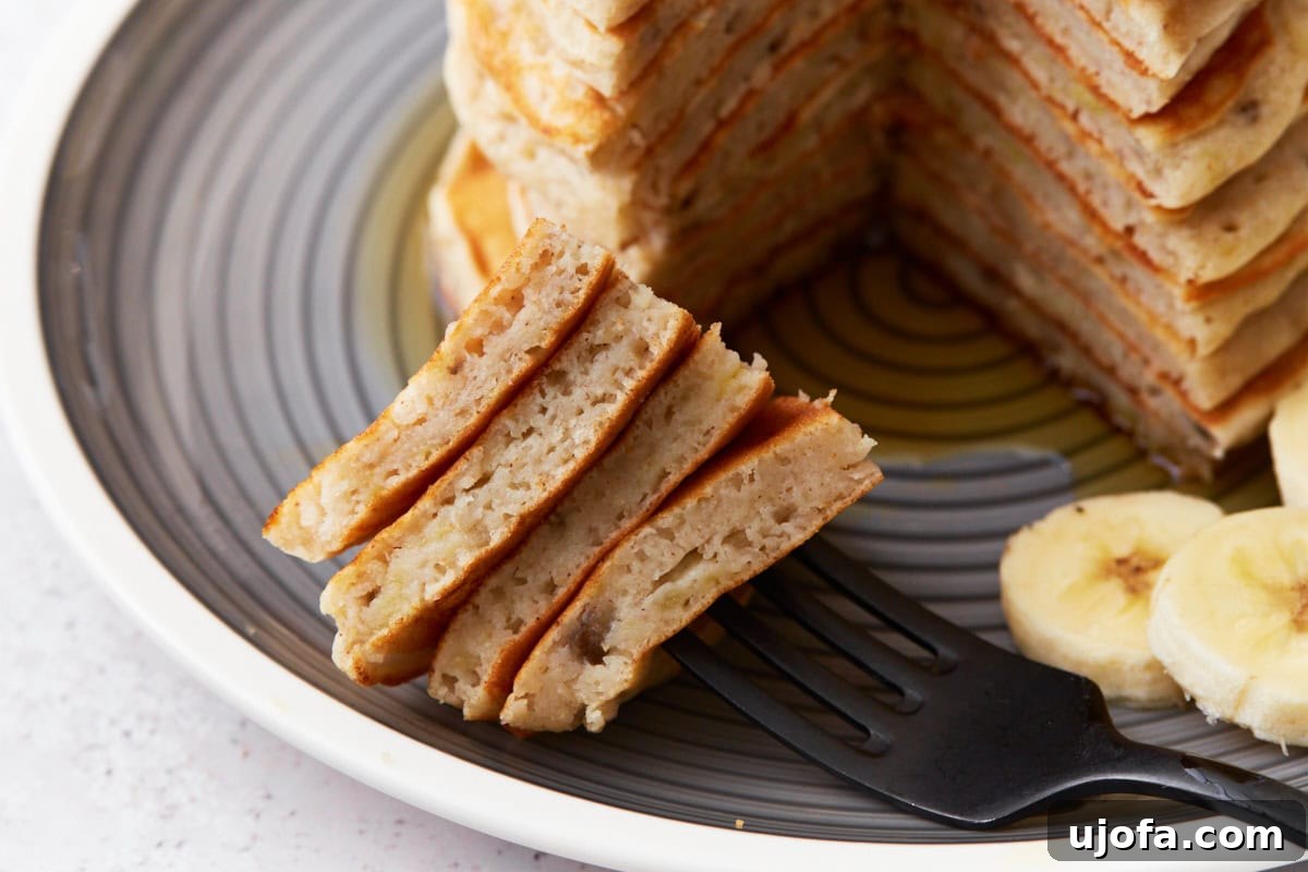 A close-up of a fork holding a stack of perfectly cut slices from fluffy gluten-free banana pancakes, revealing their tender texture. The background provides a soft blur of the remaining pancake stack and artfully arranged banana slices, emphasizing the deliciousness.