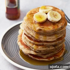 A stack of gluten-free banana pancakes on a gray plate, topped with banana slices and drizzled with maple syrup. A bottle of syrup is blurred in the background.