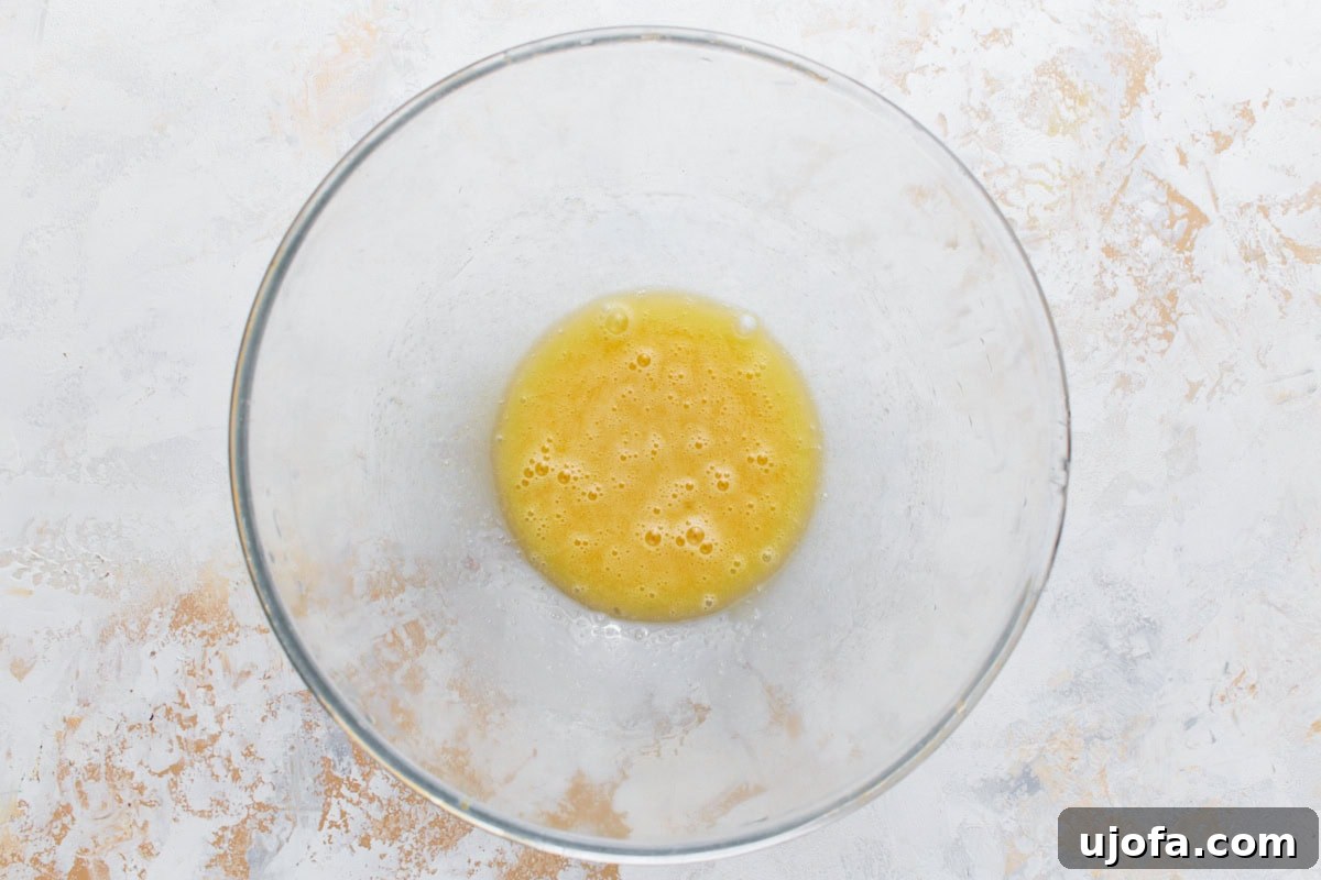 A pristine clear glass mixing bowl holds a perfectly whisked mixture of wet ingredients—egg, granulated sugar, pure vanilla extract, and vegetable oil—for gluten-free banana pancakes, creating a smooth and golden blend.
