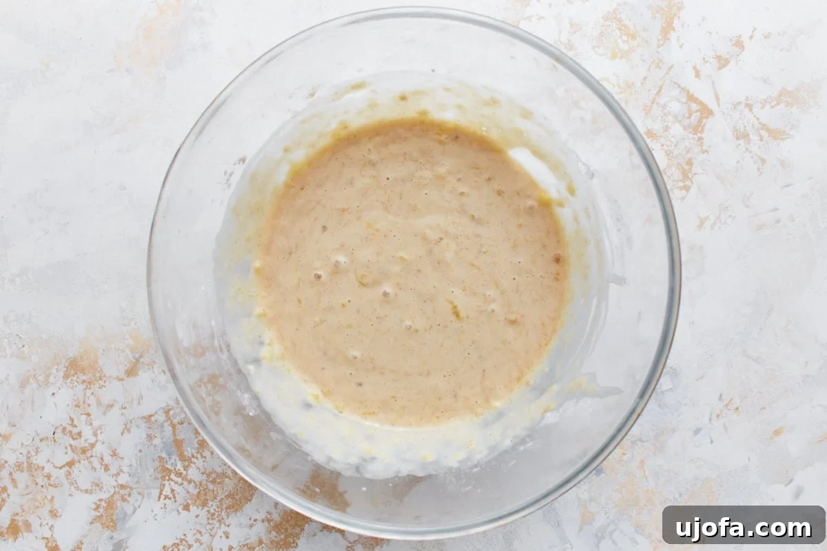 A delicious smooth pancake batter for gluten-free banana pancakes, now gently studded with visible mashed banana, all swirled together in a clear glass mixing bowl.