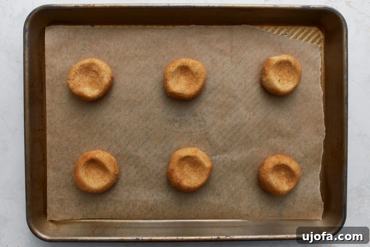 Pillowy Soft Gluten-Free Snickerdoodles 11 A baking sheet lined with parchment paper, featuring six evenly spaced, cinnamon-sugar coated gluten-free snickerdoodle dough balls, each lightly pressed in the center and ready for baking.