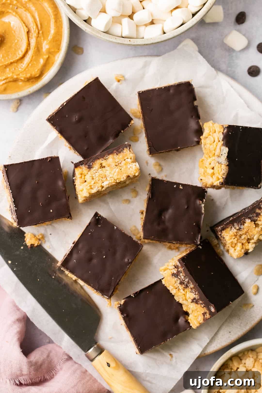 Decadent chocolate peanut butter rice krispie treats arranged on parchment paper, with a bowl of peanut butter and marshmallows in the background, ready for enjoyment.