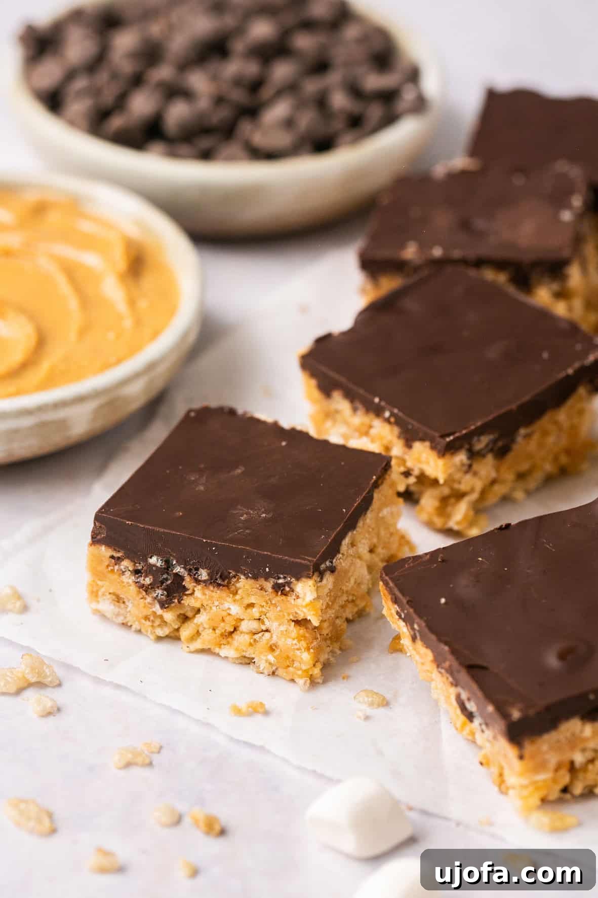 Chocolate peanut butter rice krispie treats on parchment paper, with scattered chocolate chips and a spoon scooping peanut butter in the background.