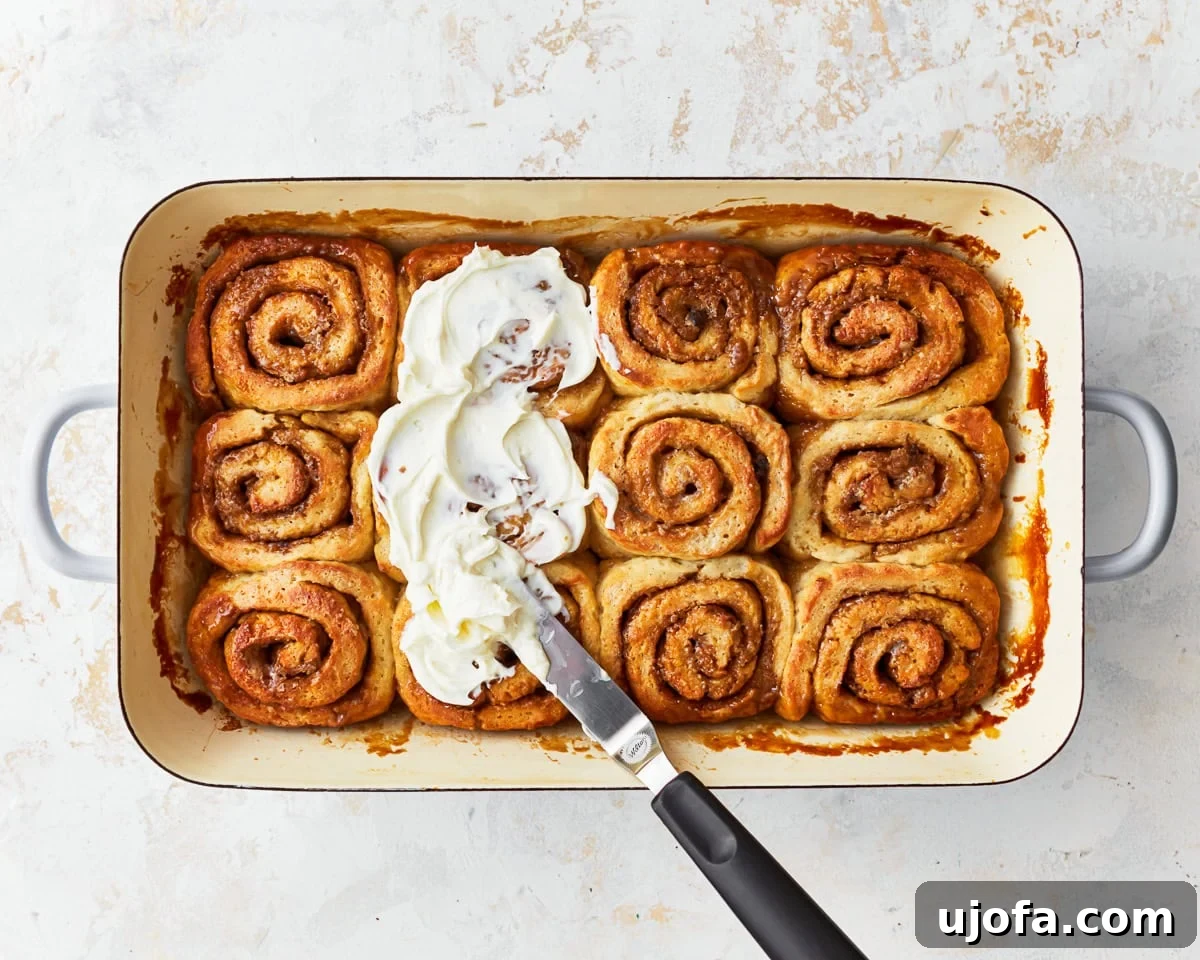 Irresistible Gluten-Free Cinnamon Buns 21 An offset spatula spreading a thick layer of cream cheese frosting over warm gluten-free cinnamon rolls in a baking dish.
