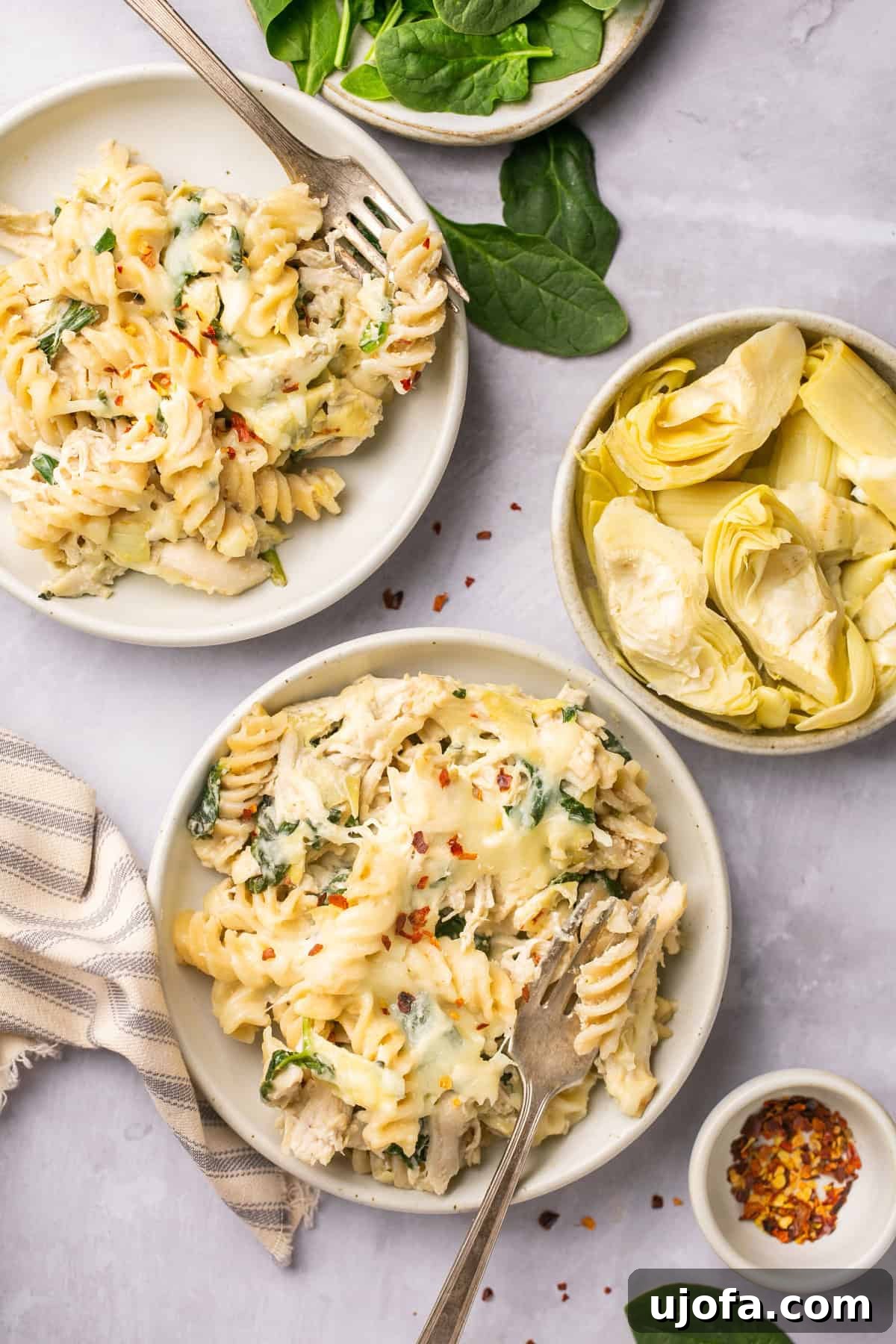 two plates filled with spinach artichoke chicken casserole next to a bowl of artichoke hearts, ready to serve.