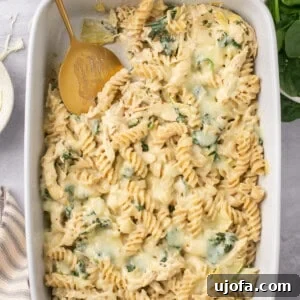 spinach artichoke chicken pasta bake in a casserole dish with a serving spoon taking a scoop out