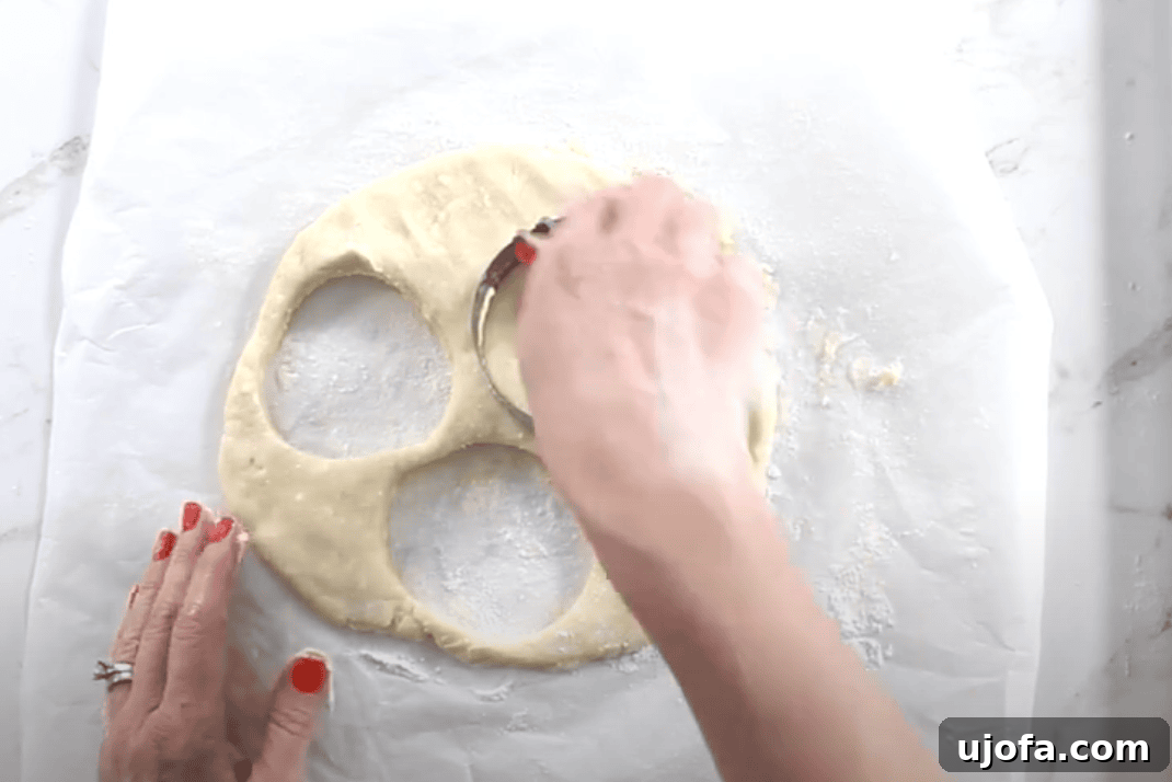 A hand presses a round biscuit cutter into rolled-out Danish dough to cut clean circles, with several rounds already removed from the dough on a floured sheet of parchment paper.